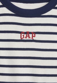 Biela rebrovaná látka s námorníckymi vodorovnými prúžkami a námorníckym golierom, s červenou vyšívanou nápisom "GAP" uprostred na hrudi.