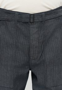 Plano detallado de pantalones de espiga gris oscuro con cinturón de tela a juego y hebilla rectangular negra, combinados con una camisa blanca.