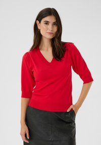 Punainen neule V-kauluksella, jossa on lyhyet hihat ja istuva vyötärö. Pehmeä rakenne, sileä kangas, yhdistetty mustan nahkahameen kanssa.