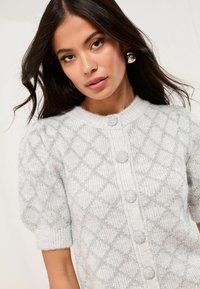 Svetlo šedý pletený kardigán s diamantom, krátkymi puff rukávmi a tromi guľatými gombíkmi s jemnou textúrovanou povrchovou úpravou.