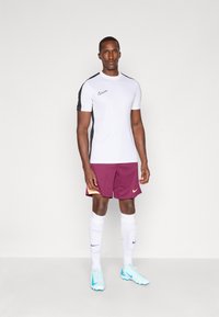 Bílá Nike tričko s černými rukávy, kaštanové šortky s oranžovými akcenty, bílé ponožky a světle modré kopačky. Jednoduchý sportovní design.
