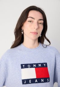 Lichtblauwe gebreide trui met een rechthoekig logo in marineblauw, rood en wit met de tekst "TOMMY JEANS"; ronde halslijn en geribbelde manchetten.