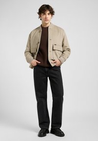 Jongeman in een beige bomberjack over een bruine sweater, zwarte jeans en zwarte schoenen, staande tegen een neutrale grijze achtergrond.