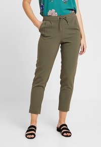 Pantalons vert olive avec taille à cordon, coupe fuselée, plis à l'avant et ourlet à la cheville ; accompagnés d'un haut floral et de sandales noires.