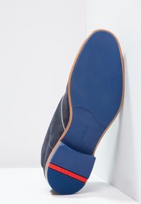Chaussure en suède gris pour homme avec une semelle en caoutchouc bleue, présentant un talon en cuir marron, des coutures blanches et une accent rouge sur le talon.