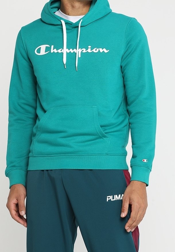 Felpa teal con logo bianco "Champion", cappuccio con cordino, tasca frontale e polsini a costine. Indossata con pantaloni verde scuro con logo.