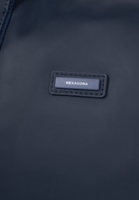 Materiale testurizzato blu navy con un'etichetta nera rettangolare in rilievo con la scritta "HEXAGONA" in caratteri argentati. Dettaglio della cerniera visibile.