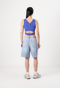 Top court bleu sans manches avec découpe au dos et détail de boutons, associé à un short Bermuda en denim clair. Le mannequin porte des baskets grises et des chaussettes blanches.