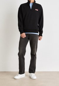 Persona che indossa un maglione nero a zip con logo rosso, una camicia azzurra chiara sotto, jeans scuri e sneakers bianche, che sta in piedi su un pavimento beige.