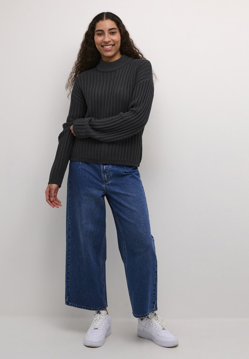 Ciemnoszara sweter prążkowany z wysokim kołnierzem, w zestawie z szerokimi niebieskimi dżinsami i białymi trampkami. Gładka struktura dzianiny, luźny krój.
