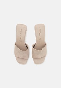 Beige semskede åpen tå mules med en bred krysset stropp og firkantet hæl, med en glatt tekstur og subtil merking på innersålen.