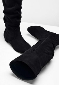 Bottes en daim noires au dessus du genou avec une semelle plate ; design décontracté avec détail de fermeture éclair sur le côté ; texture lisse avec une finition douce.