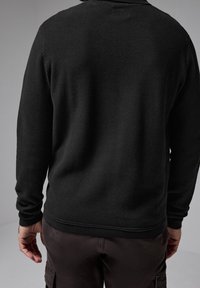 Schwarzer Strickpullover mit strukturierter Oberfläche, langen Ärmeln sowie gerippten Bündchen und Saum, kombiniert mit dunklen Hosen, von hinten gezeigt.