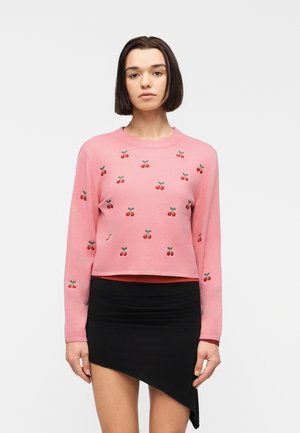 Kobieta o krótkich ciemnych włosach, ubrana w różowy sweter z wzorem wiśni oraz asymetryczną czarną spódnicę, stojąca na tle jednolitego białego tła.