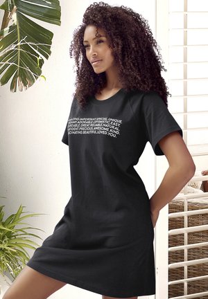 Frau mit lockigem Haar, die ein schwarzes Kleid mit weißen inspirierenden Worten trägt, steht nahe bei großen blätterreichen Pflanzen und einem Fenster mit Jalousien.