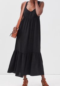 Femme portant une robe maxi noire sans manches avec un ourlet à volants, un sac bandoulière marron, et des sandales à talons hauts marron avec des lanières.