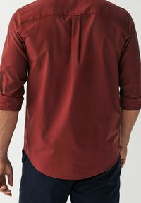 Camisa burdeos de manga larga con puños remangados, con un pliegue en la espalda y dobladillo redondeado. Fabricada con tela suave y un ligero brillo sutil.