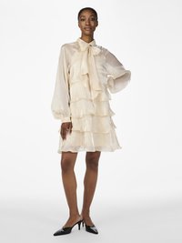Vrouw staat met haar hand op de heup, gekleed in een crèmekleurige, gelaagde chiffon jurk met lange mouwen en een striknek, gecombineerd met zwarte kitten heel muiltjes.