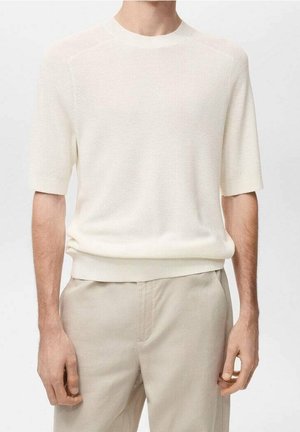 Man wearing a light beige short-sleeve knit sweater tucked into matching beige trousers on a plain background.