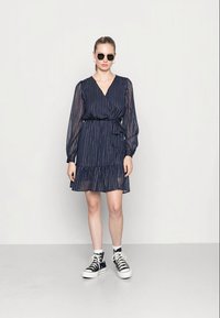 Vestido azul-marinho de mangas longas com riscas brancas verticais, decote em V, cinto na cintura e bainha com babados; combinado com ténis pretos de cano alto e óculos de sol.