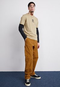 T-shirt di cotone beige con logo nero, indossata sopra una maglietta nera a maniche lunghe, abbinata a pantaloni in velluto a coste marroni e sneakers nere.