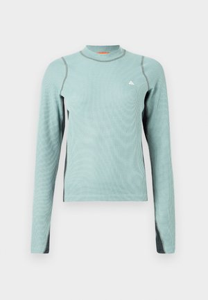 Lichtgroene blouse met lange mouwen en hoge hals, met grijze accenten en een textuurstof. Klein logo op de borst.