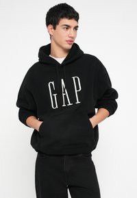 Mladý muž s krátkymi tmavými vlasmi, oblečený v čiernej mikine GAP s kapucňou a čiernych nohaviciach, ruky v vreckách mikiny, pozerajúci sa bokom na bielom pozadí.