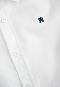 Biała koszula z długim rękawem, wykonana z gładkiego materiału, z małym, granatowym haftem lwa oraz standardowym zapięciem na guziki.