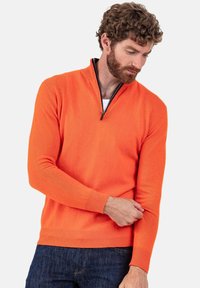 Pull-over orange avec un col demi-fermeture éclair, poignets et ourlet côtelés ; fabriqué en tissu tricoté doux. Porté par-dessus une chemise blanche avec un jean foncé.