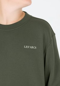 Olivegroene sweatshirt met een geribde ronde hals en lichte schouderdetails, met "LES'ARCS" in het wit geborduurd. Zachte textuur.