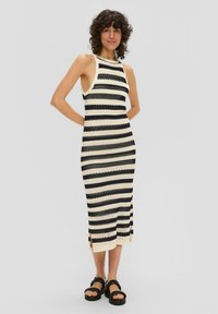 Robe en tricot avec des rayures noires et crème horizontales, sans manches, fentes sur les côtés et une silhouette ajustée. Associée à des sandales noires à semelles épaisses.