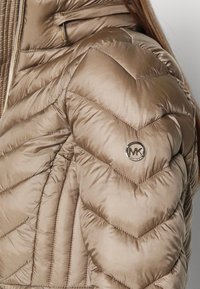 Beige Steppjacke mit Puffer-Effekt, Schulterbereich mit sichtbarem Michael Kors Metalllogo und Reißverschlussdetails nahe dem Kragen.