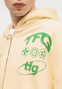 Κοντινό πλάνο ενός ατόμου που φοράει ανοιχτό κίτρινο φούτερ με φερμουάρ και πράσινα αφηρημένα σύμβολα και γράμματα "tfg" στον ώμο.