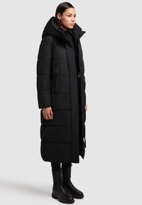 Palton puffer lung, negru, cu guler înalt, buzunare laterale cu fermoar, design matlasat și glugă. Modelul poartă cizme negre.