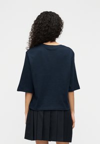 Μπλε σκούρο oversized μπλουζάκι από μαλακό ύφασμα με στρογγυλή λαιμόκοψη και κοντά μανίκια, συνδυασμένο με μαύρη πλισέ φούστα.