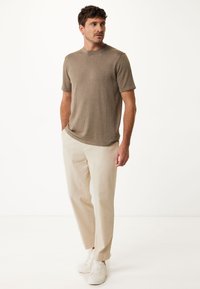Maglietta a maniche corte in maglia marrone, con scollatura rotonda, abbinata a pantaloni di lino beige chiaro e sneakers bianche, in piedi su uno sfondo semplice.