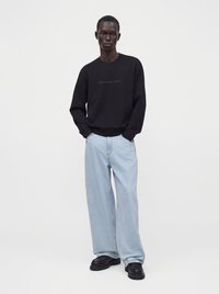 Homme portant une chemise noire à manches longues avec texte, jean large bleu clair et chaussures noires, debout les mains dans les poches.