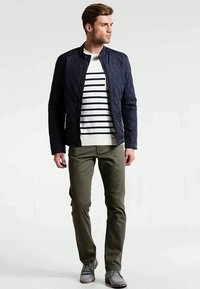 Jovem em pé, vestindo uma jaqueta acolchoada navy, camisa listrada em branco e navy, calças oliva e sapatos cinza, olhando para o lado.