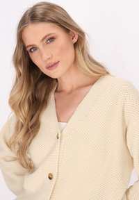 Světle krémový pletený cardigan s výstřihem do V, který zdobí dva dřevěné knoflíky a strukturovaný vzor. Světlé rukávy a uvolněný střih.