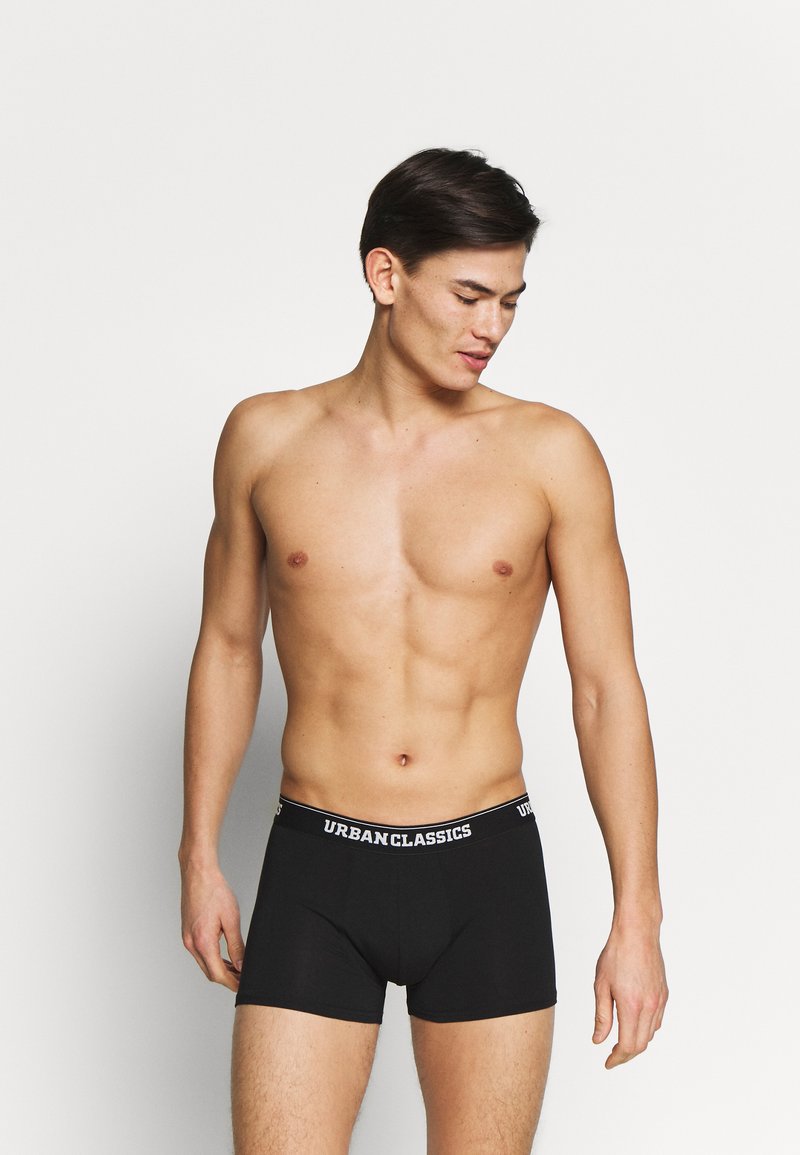 Schwarze Boxershorts aus dehnbarem Stoff mit elastischem Bund, auf dem das Logo "URBANCLASSICS" in Weiß prangt. Glatte Textur, figurbetonter Schnitt.