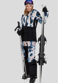 Vrouw in een ski-jas en broek met patroon houdt skistokken en ski's vast, draagt zwarte handschoenen, skischoenen, een muts en een skibril op haar hoofd.