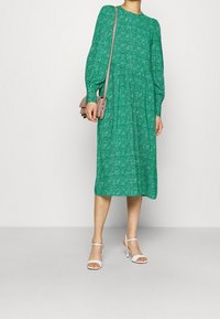 Abito verde a fantasia con maniche lunghe a sbuffo, scollatura rotonda e gonna svasata, abbinato a una borsa a tracolla rosa chiaro e sandali bianchi.
