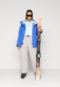 Μπλε σκιών jacket, ανοιχτό μπλε μπλούζα, γκρίζες παντελόνες σκι, μαύρες μπότες σκι. Κρατώντας καφέ σκι με μαύρους συνδέσμους. Γυαλιά ηλίου. Επίπεδο υπόβαθρο.