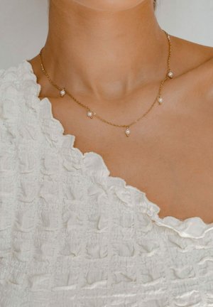 Goldene Kettenhalskette mit fünf gleichmäßig verteilten kleinen runden Perlen. Haut ist unter der Kette sichtbar, dazu ein strukturierter weißer Stoffoberteil.