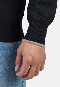 Schwarzer Strickpullover mit gerippter Textur, der kontrastierende graue Akzente an den Bündchen aufweist. Die Hand ruht auf hellblauem Denim.
