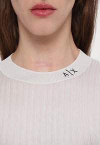 Viegls ribu adījuma topiņš off-white krāsā ar vieglu spīdumu. Iezīmēts ar pieguļošu siluetu un apaļu apkakli, ar "AIX" zīmolu.