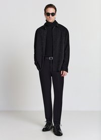 Camicia nera con trama sopra un dolcevita nero, abbinata a pantaloni neri e scarpe stringate. Accessori includono occhiali da sole neri.