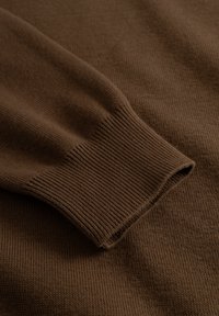 Pull marron en maille avec poignets côtelés, présentant une texture douce et une surface lisse. Le tissu semble épais et chaud, mettant l'accent sur le confort.