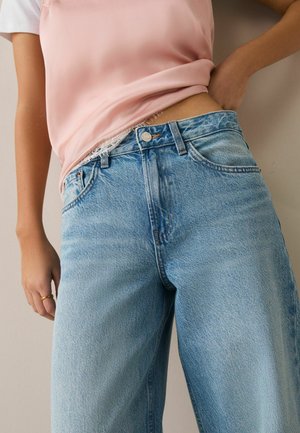 Nő világoskék, magas derekú, bő farmernadrágot és rózsaszín selyemtornyot visel, amely fölött csipkés fehérnemű látható.