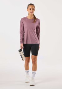 Pitkähihainen lila-urheilupaita, mustat pyöräilysortsit, valkoiset lenkkarit ja kirkas vesipullo mustalla kannella, jossa on logo.